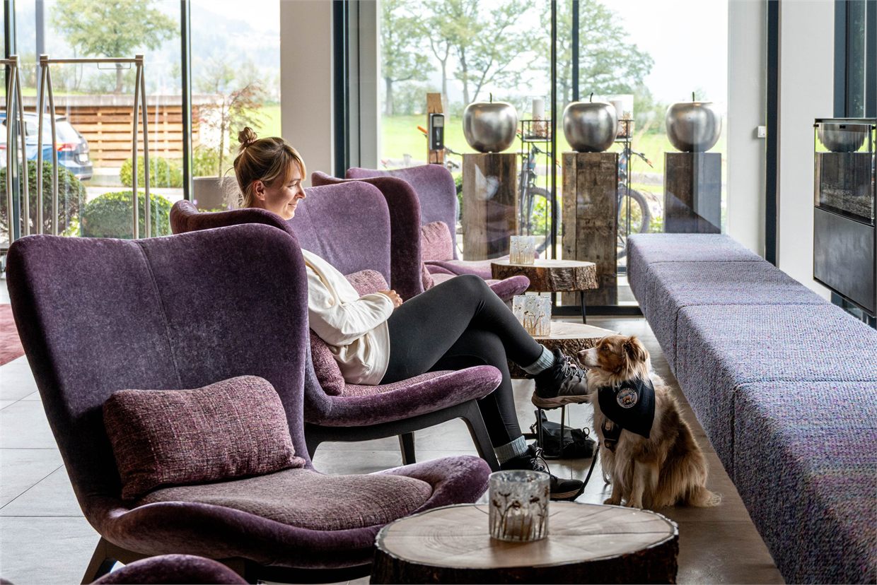 Julia Doubrawa sitzt entspannt in einem Sessel in einem hellen Loungebereich, ihr Hund wartet ruhig neben ihr